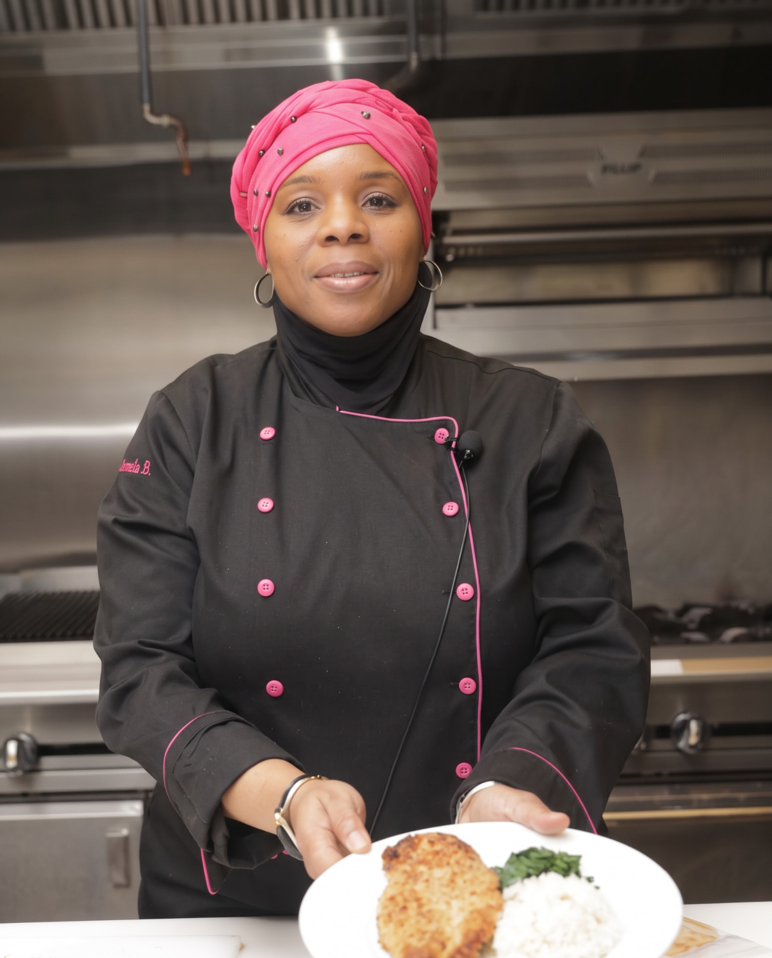 Chef Jamela B. in her black chef coat with pink trim and pink headwrap, holding a plate of breaded cutlet, rice, and greens in a professional kitchen.